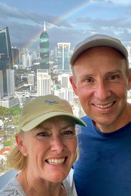 John & Bev, retirement travelers, standing on balcony with Panama City skyline in rear with rainbow