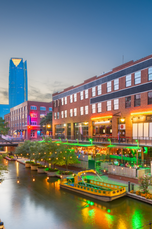 A city view of a skyscraper and river walk with restaurants along the side of it in Oklahoma City.