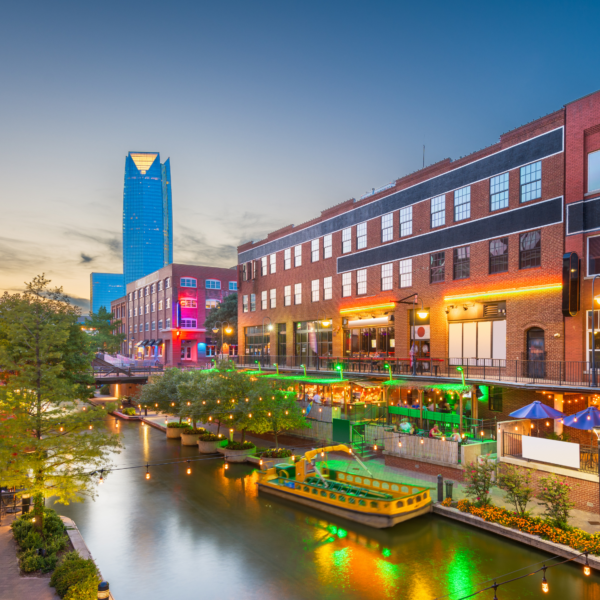 A city view of a skyscraper and river walk with restaurants along the side of it in Oklahoma City.